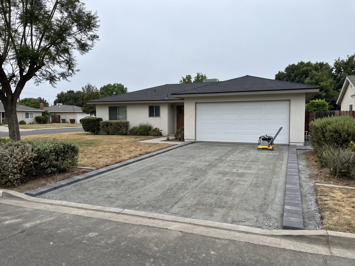 A two car driveway bed of compacted gray crushed rock with a charcoal paver border set along both edges, running from the curb to a closed garage door, with a plate compactor parked near the garage.
