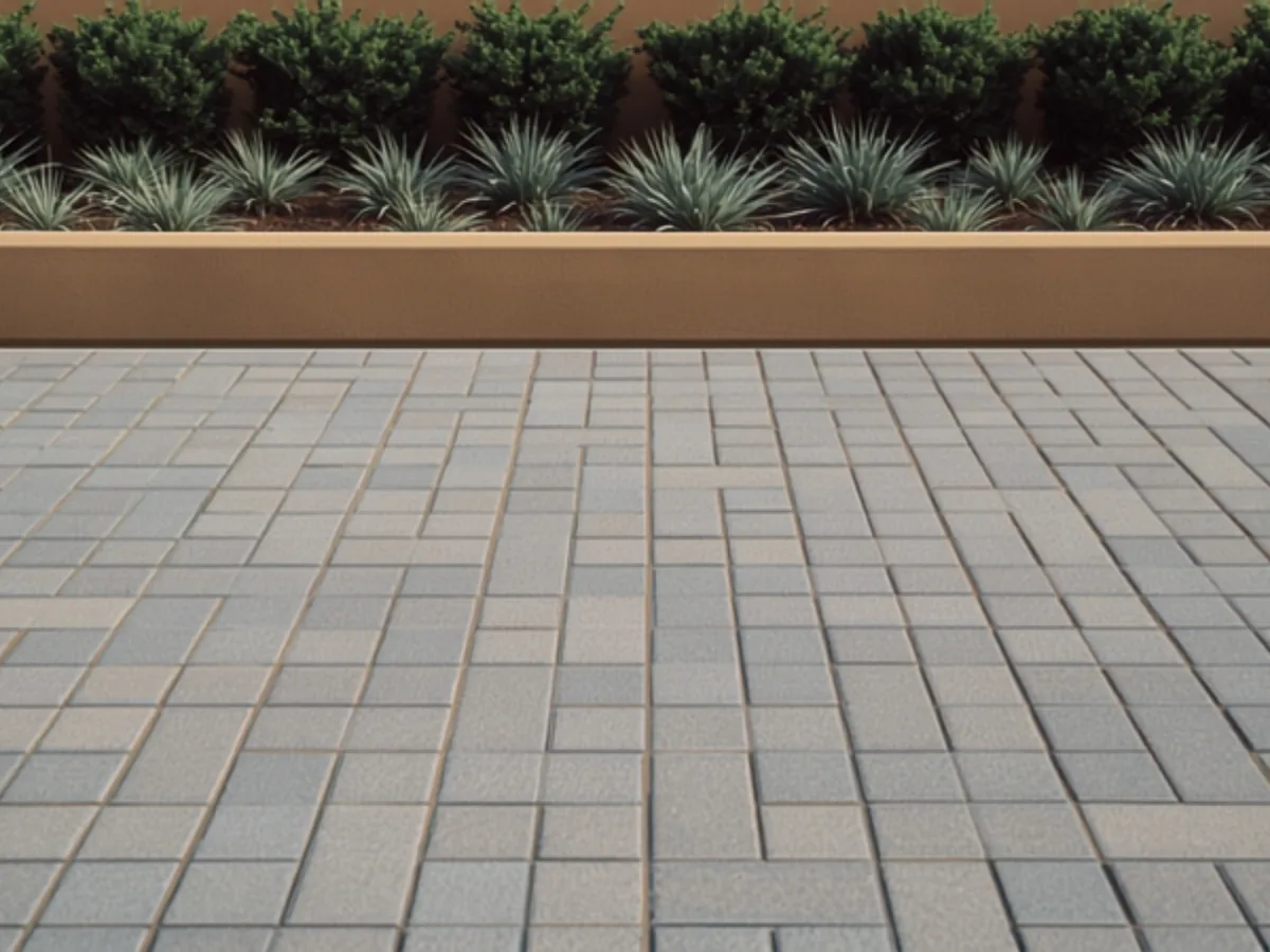 Close view of gray concrete pavers in a mixed pattern of squares and rectangles with sand filled joints, beside a raised planter of agaves and shrubs.