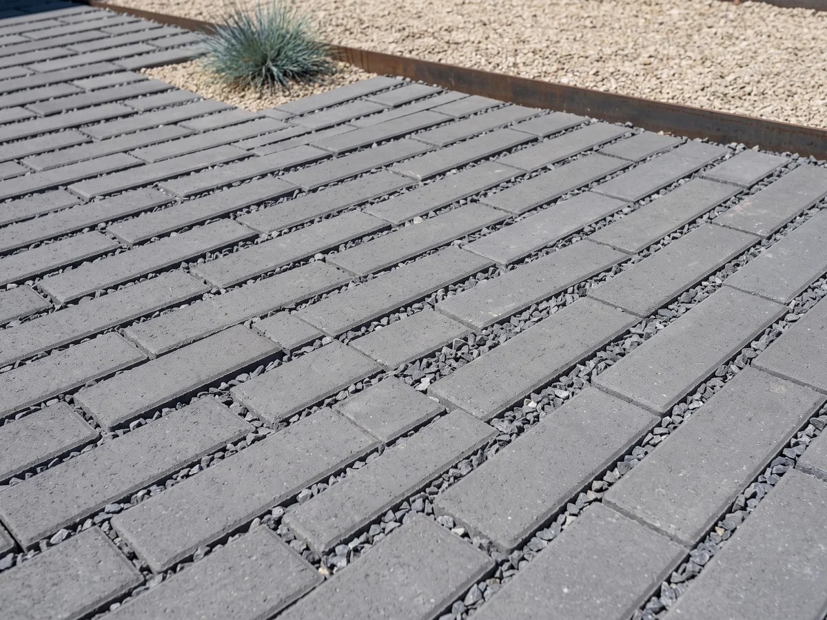 Charcoal concrete pavers in a running bond with wide open joints filled with loose crushed stone, meeting a steel edge and a gravel bed.